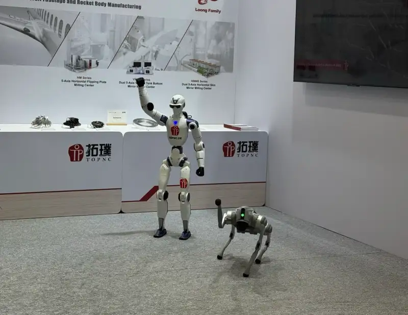 Unitree R1 zwaait op de stand van TOPNC op de AMB in Stuttgart, de Go2 Pro staat ernaast.